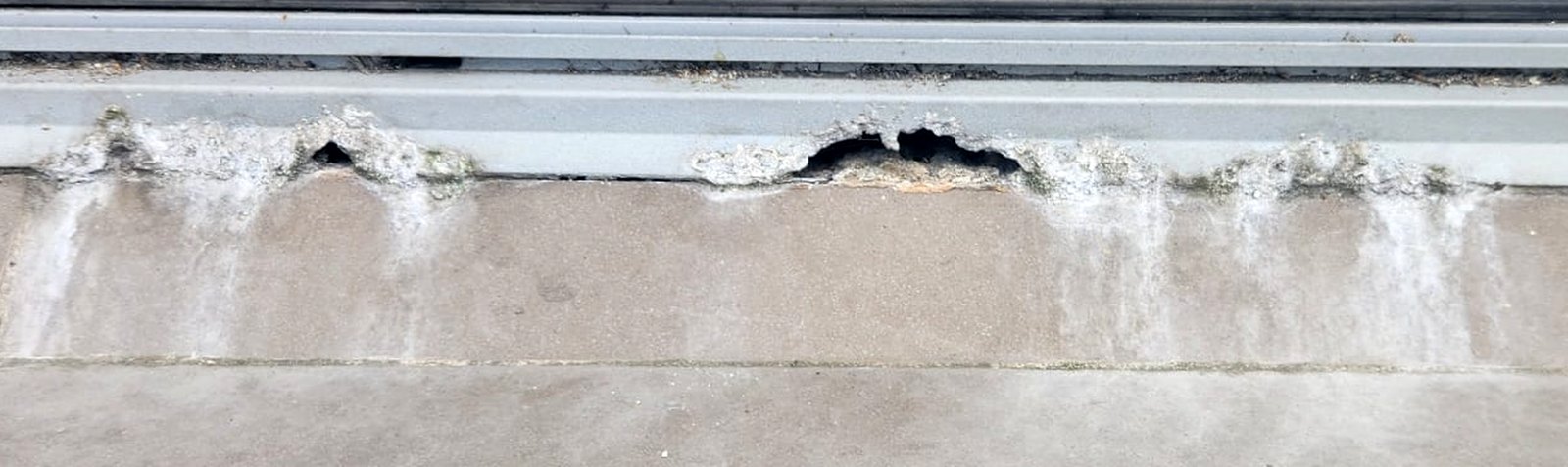 Close-up of an aluminium sliding door sill with holes corroded through its front edge and white corrosion product running down the tiles